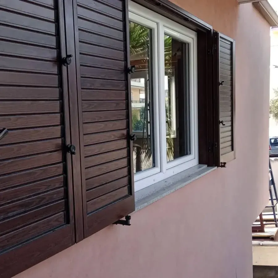 Finestra con persiane a stecche marrone scuro contro una parete in stucco rosa.