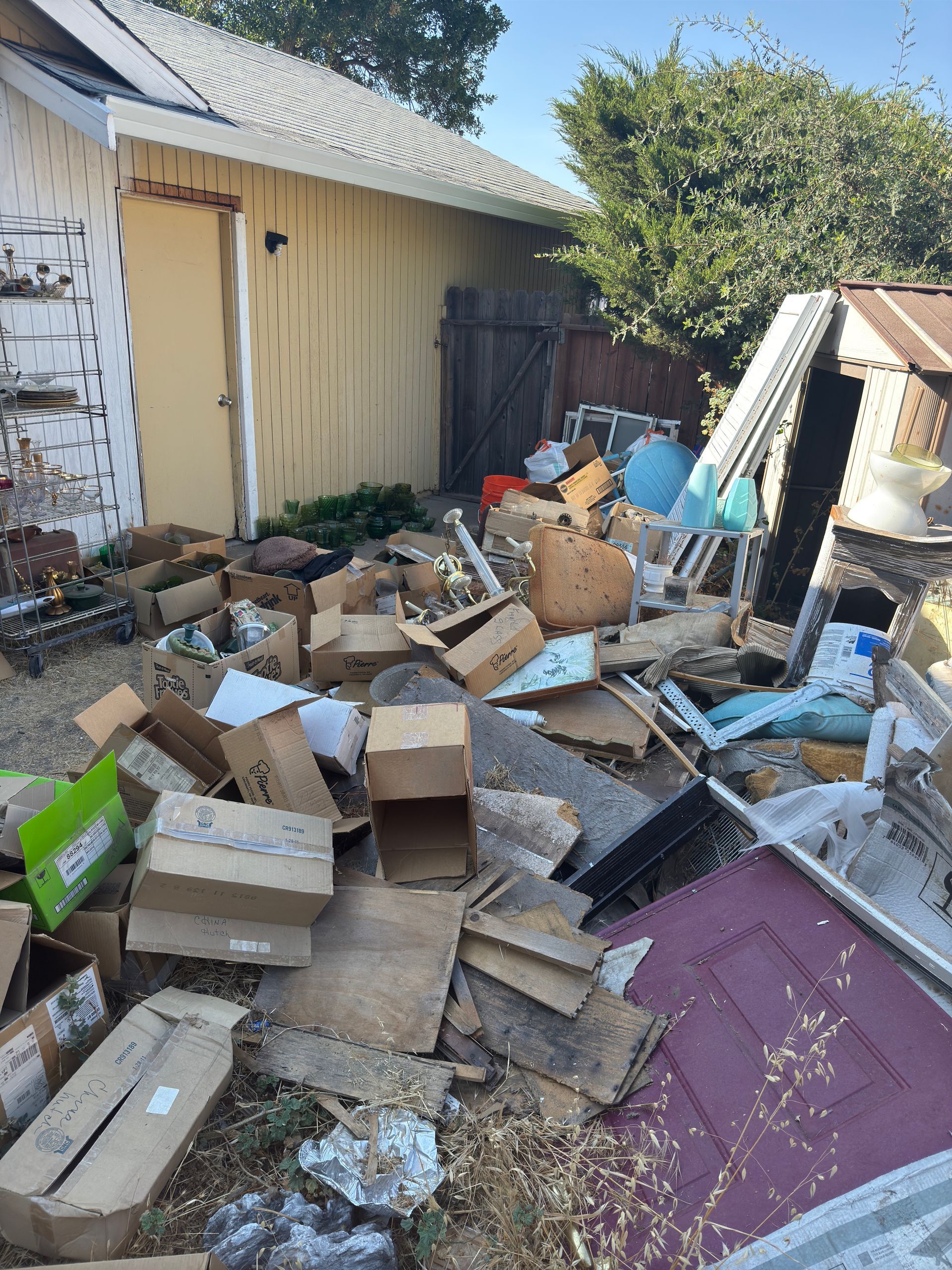 A yard cluttered with numerous discarded cardboard boxes, debris, and a purple door leaning against an outdoor structure.