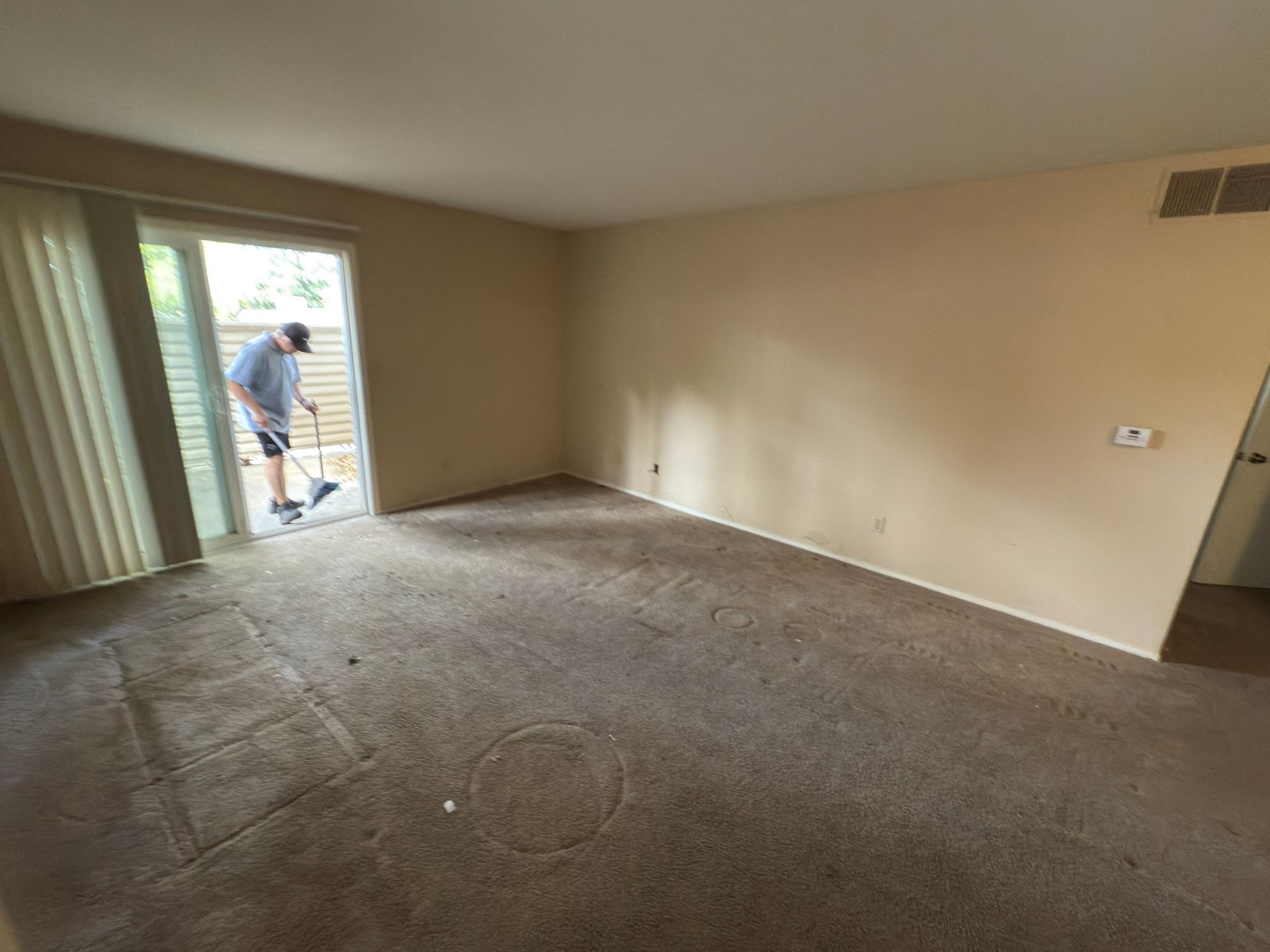 A person uses a tool to clean a tan-carpeted living room near a glass patio door.