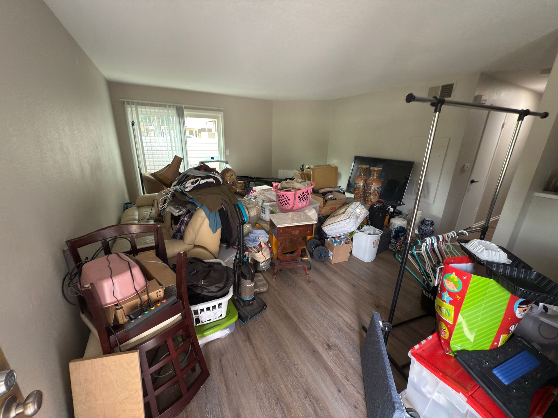 A room with light-colored flooring, cluttered with various boxes, furniture, bins, and personal belongings.