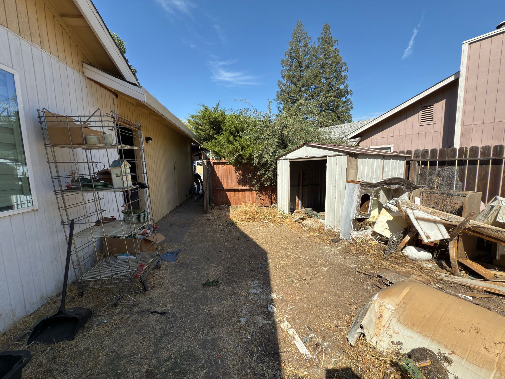 A cluttered side yard between two houses, featuring a metal shed, scattered debris, and dry, uneven ground.