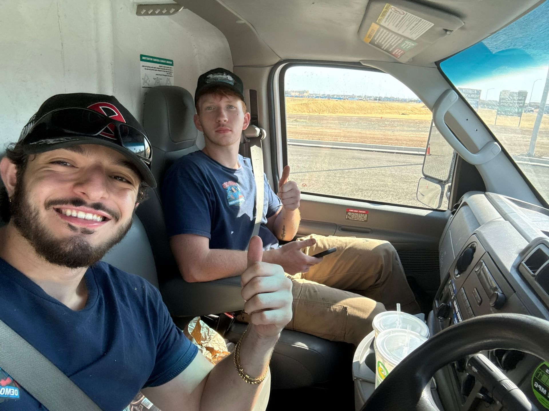 Two people in the front seats of a vehicle, smiling and giving thumbs up, with a desert landscape visible through windows.