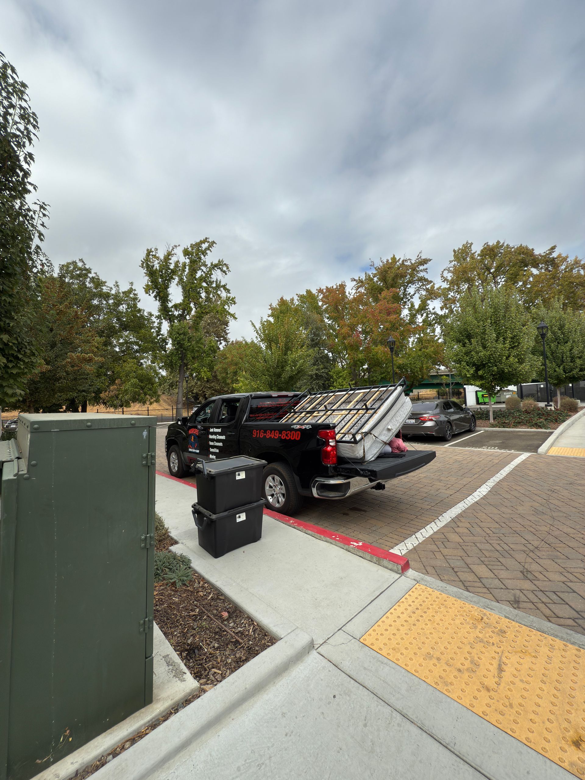 A black pickup truck parked in a lot, loaded with equipment and white containers, with two cases on the sidewalk nearby.