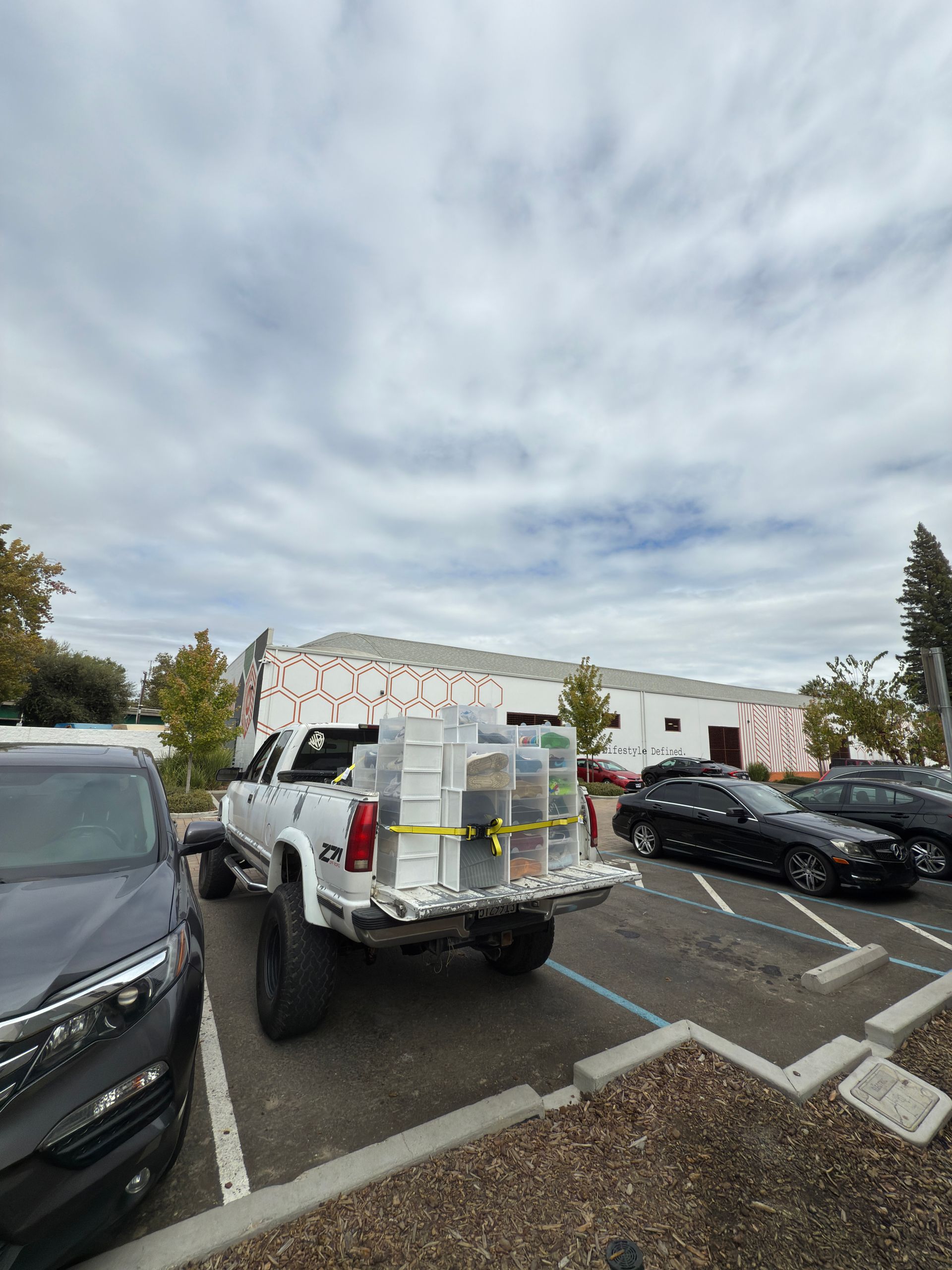 A white pickup truck parked in an outdoor lot, carrying stacked containers secured with yellow straps in the truck bed.