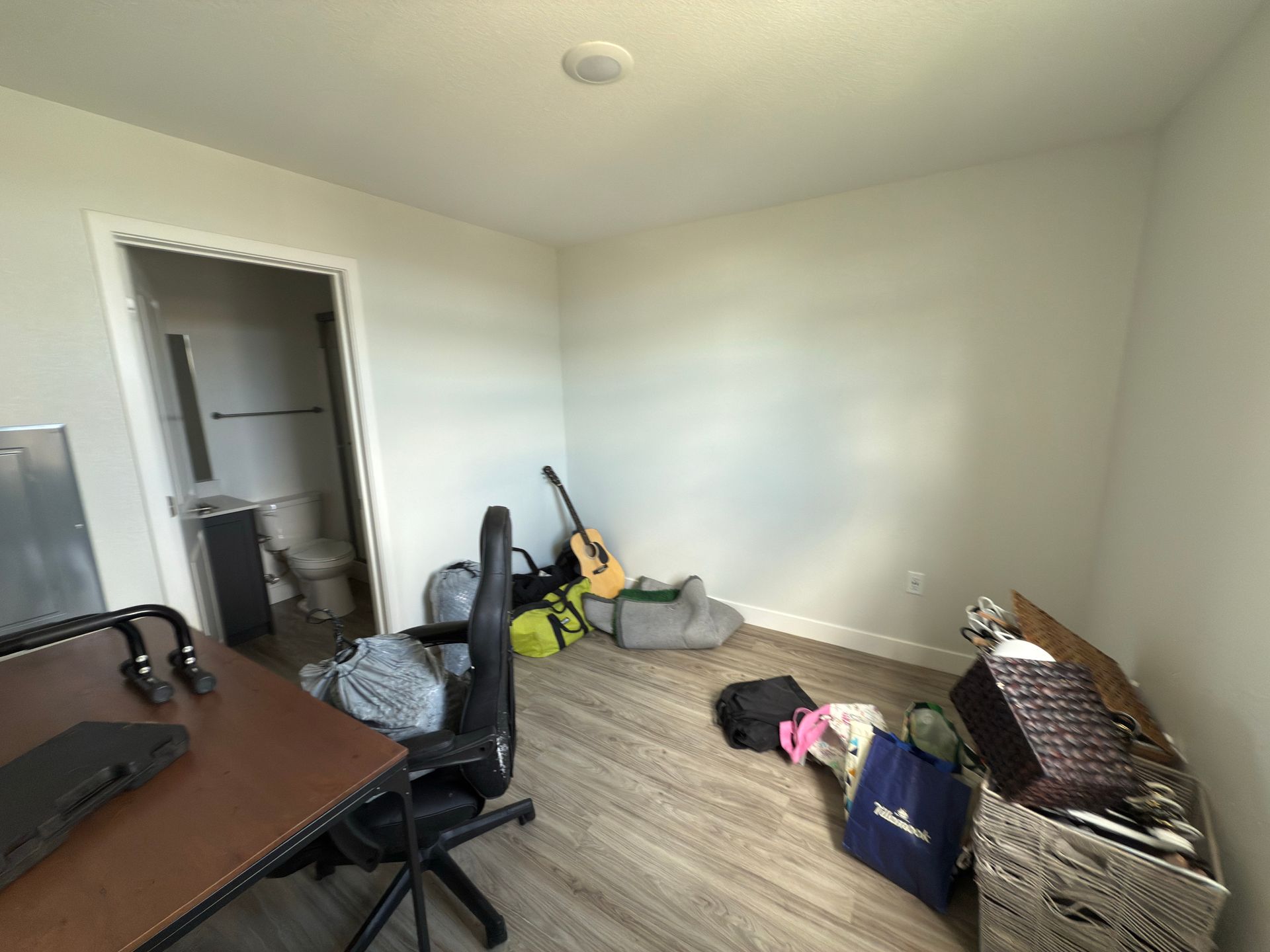A room with wood-look flooring, a desk, a black office chair, an acoustic guitar, bags, and a cardboard box.