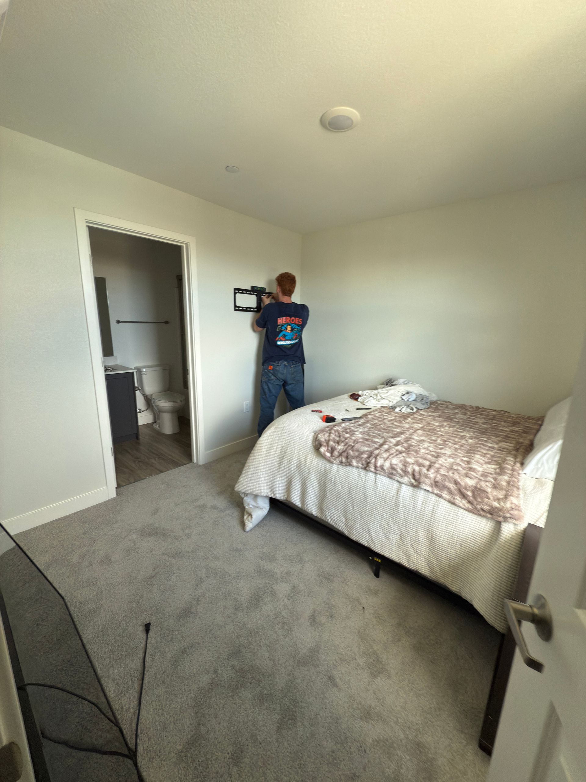 A person standing on a bed while mounting a television bracket to a bedroom wall, with a bathroom visible in the background.