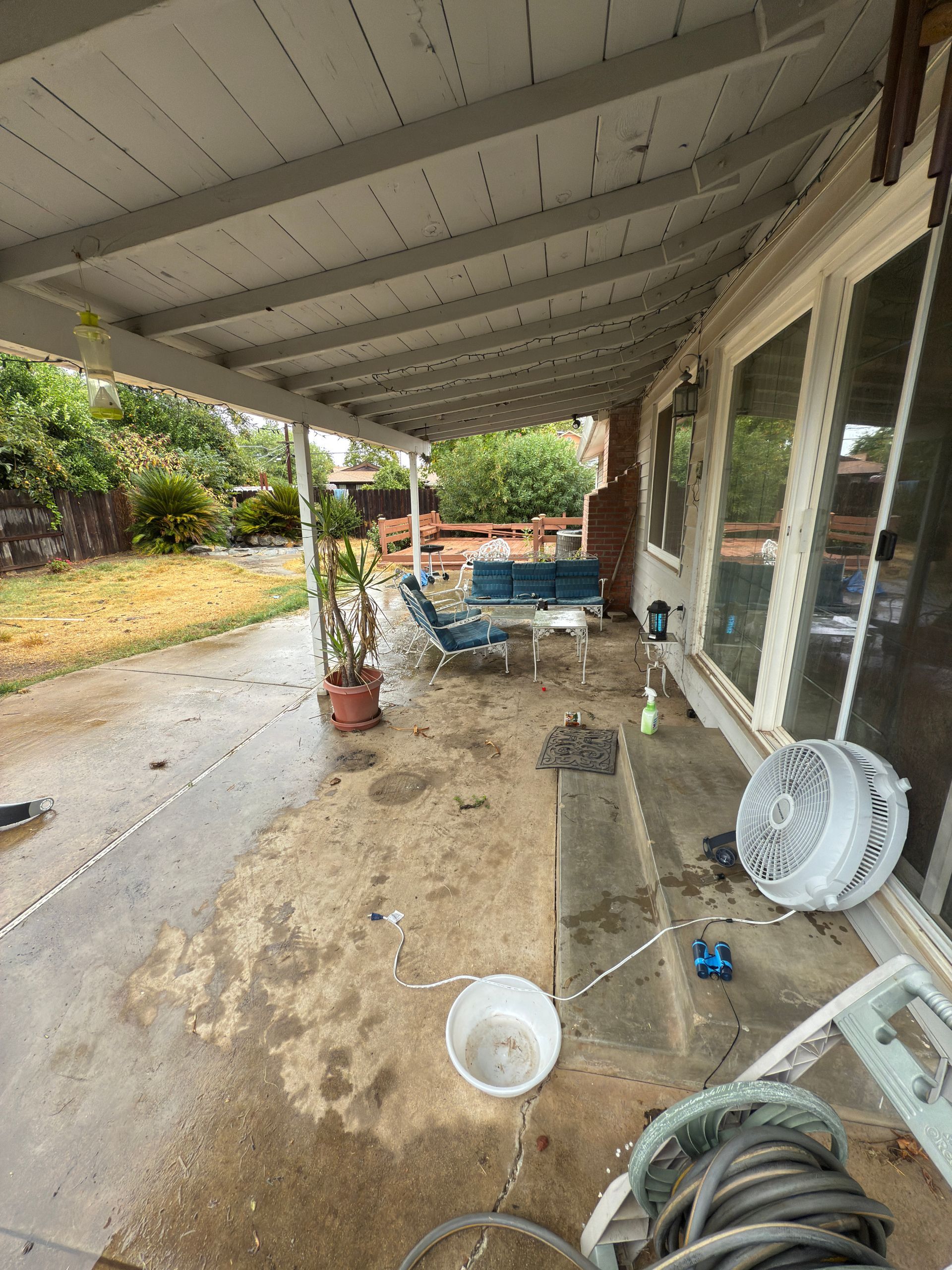 A covered patio with a concrete floor, patio furniture, a large fan, a plant in a pot, and a hose reel near a home.