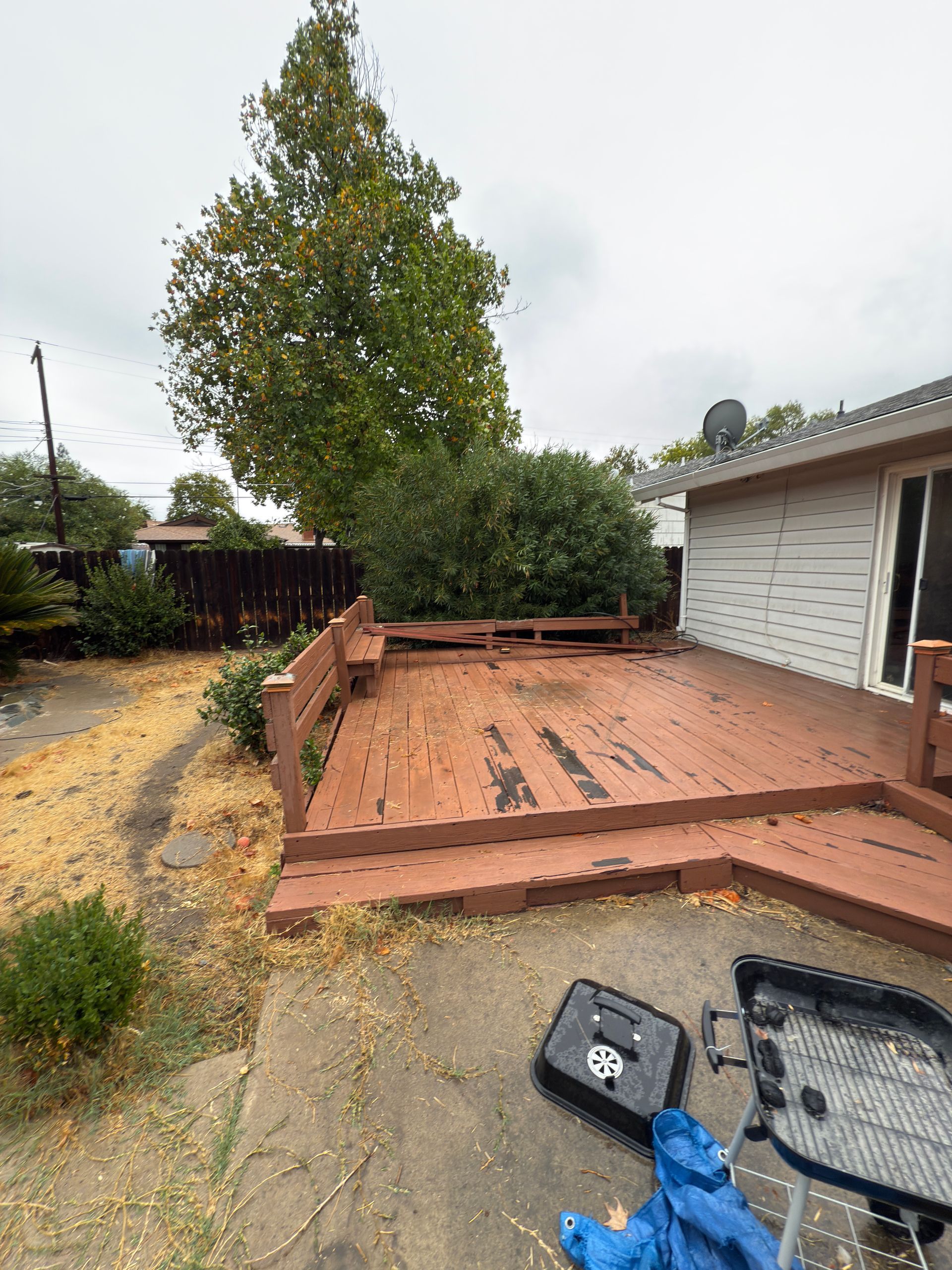 A slightly weathered, reddish-brown wooden backyard deck adjacent to a white house with a grill in the foreground.
