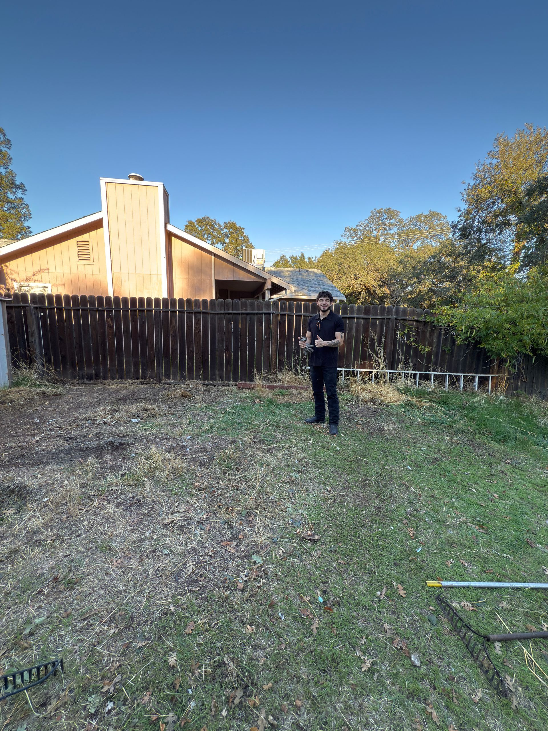 A person stands in a sparse backyard with brown grass, facing a wooden fence and a house with a prominent chimney.