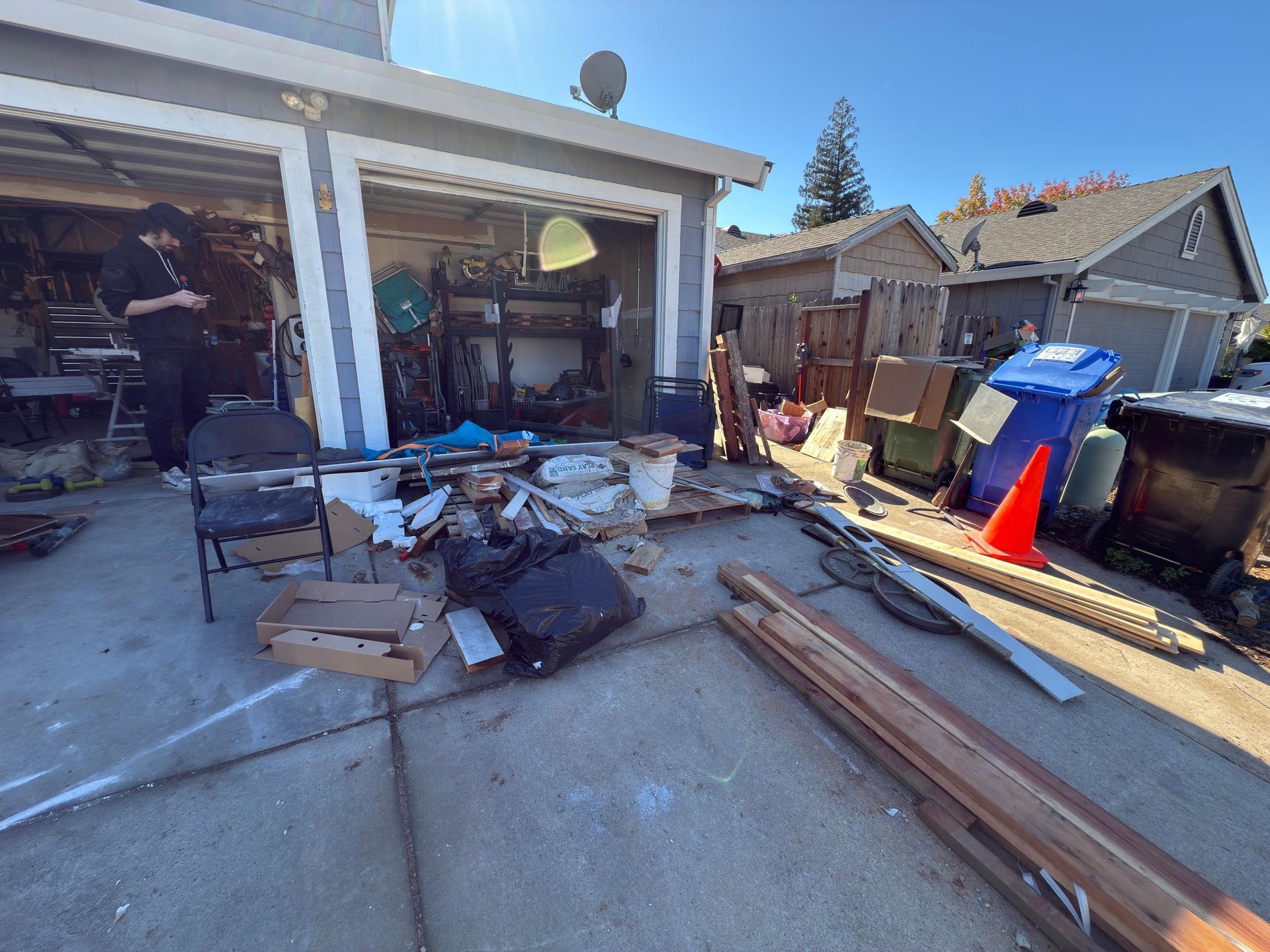 A person stands in a cluttered garage with debris and building materials scattered on the concrete driveway outside.