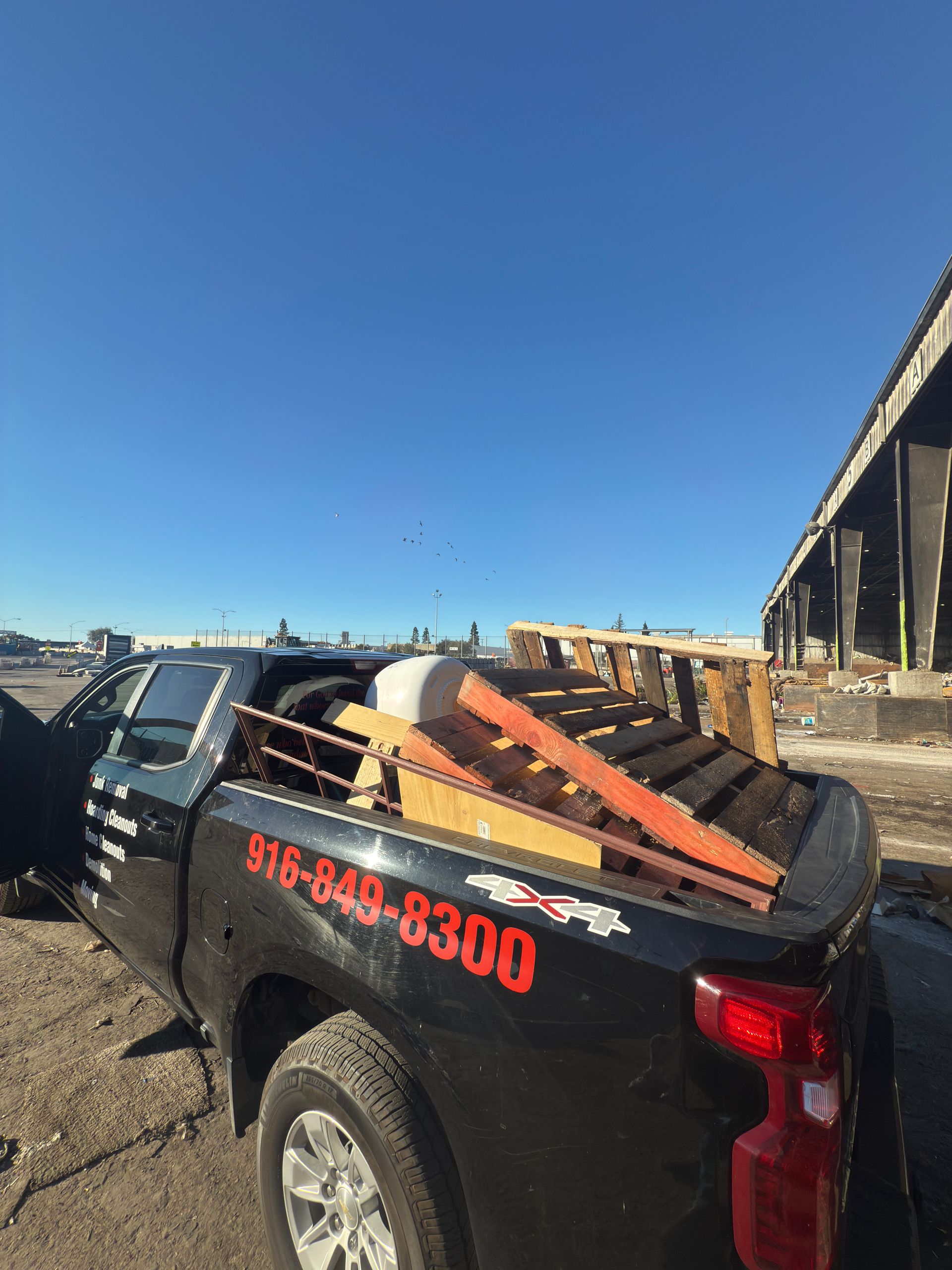 A black pickup truck loaded with wooden pallets and metal scrap sits on a dirt lot under a clear blue sky.
