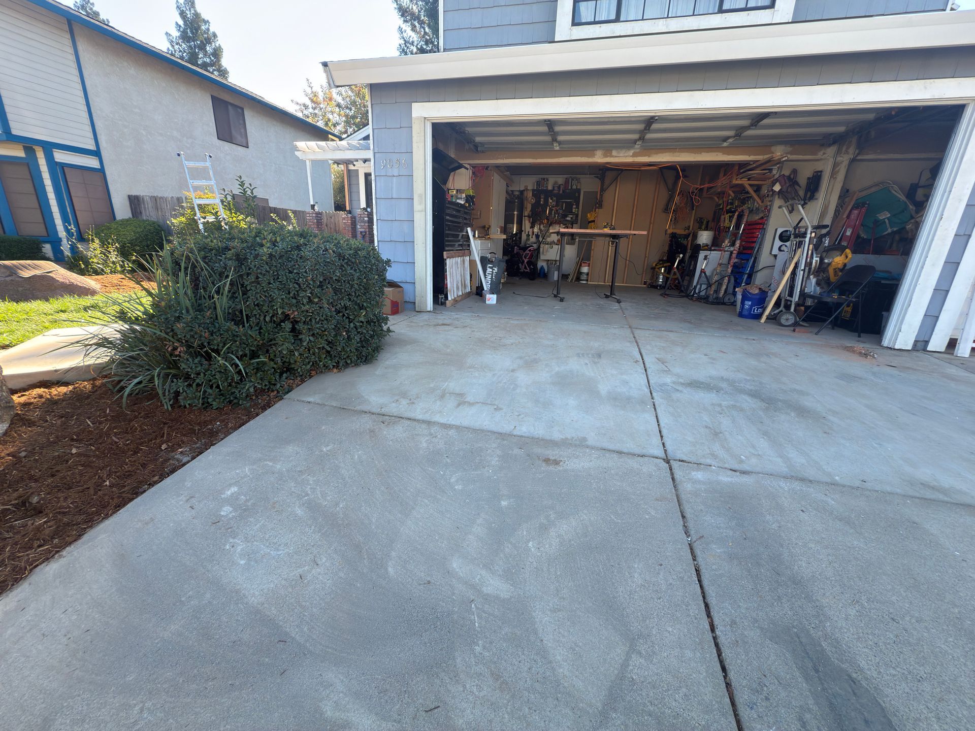 A view of a blue residential house exterior, a driveway, a large bush, and an open garage containing various items.
