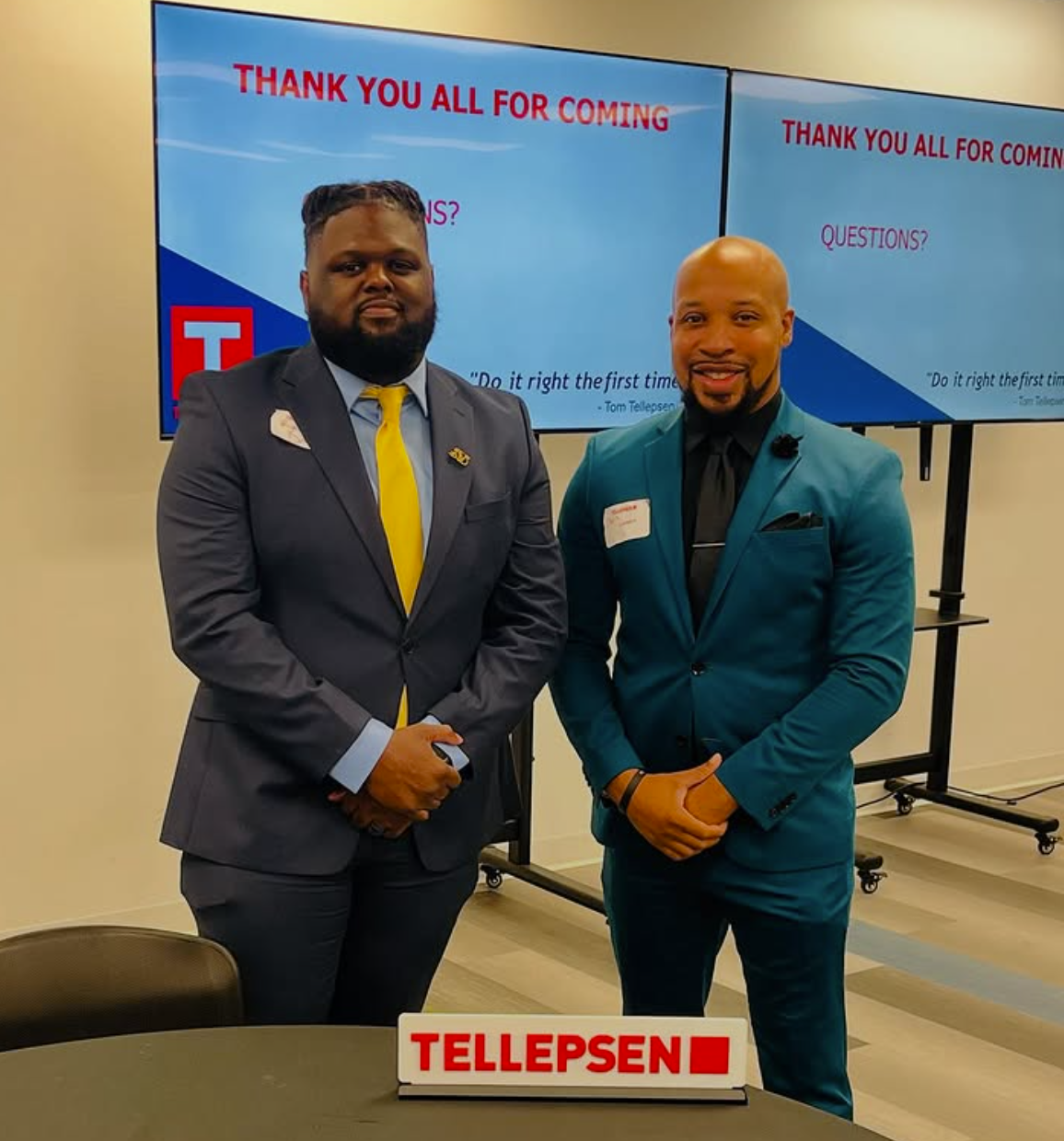 Two men in suits stand in front of screens that read