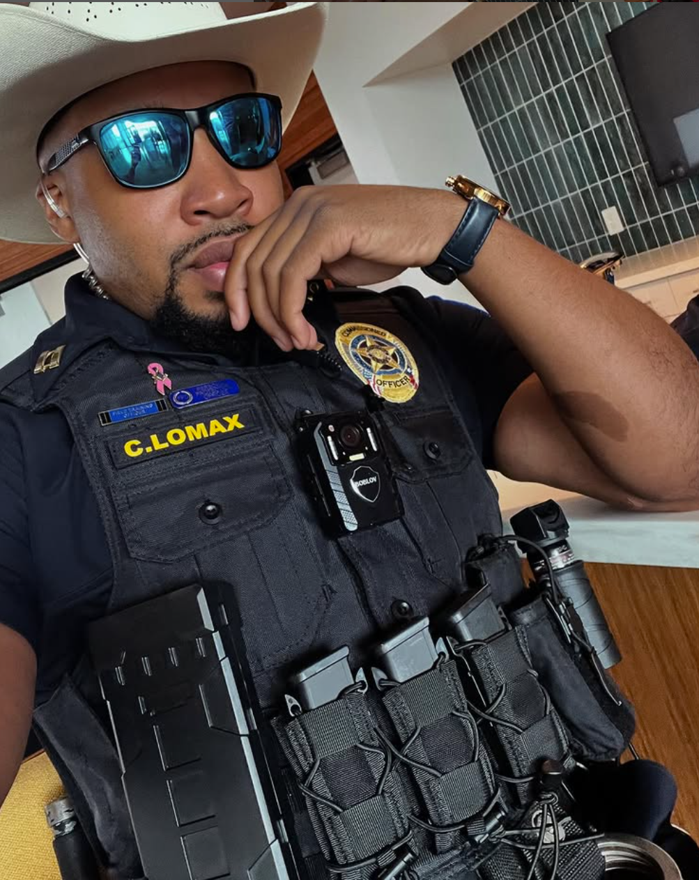 A Black police officer wearing a cowboy hat and sunglasses. He's in uniform, posing indoors.