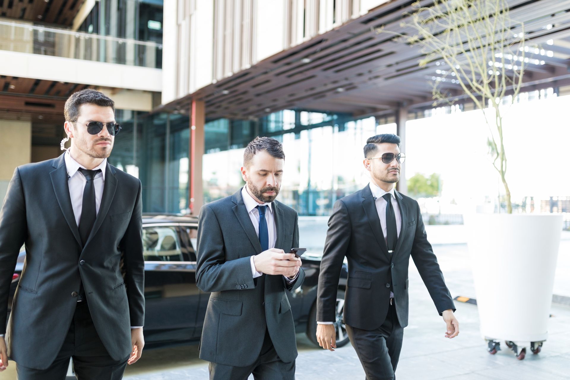 Three men in suits: one using a phone, two bodyguards with sunglasses, walking outside a modern building.