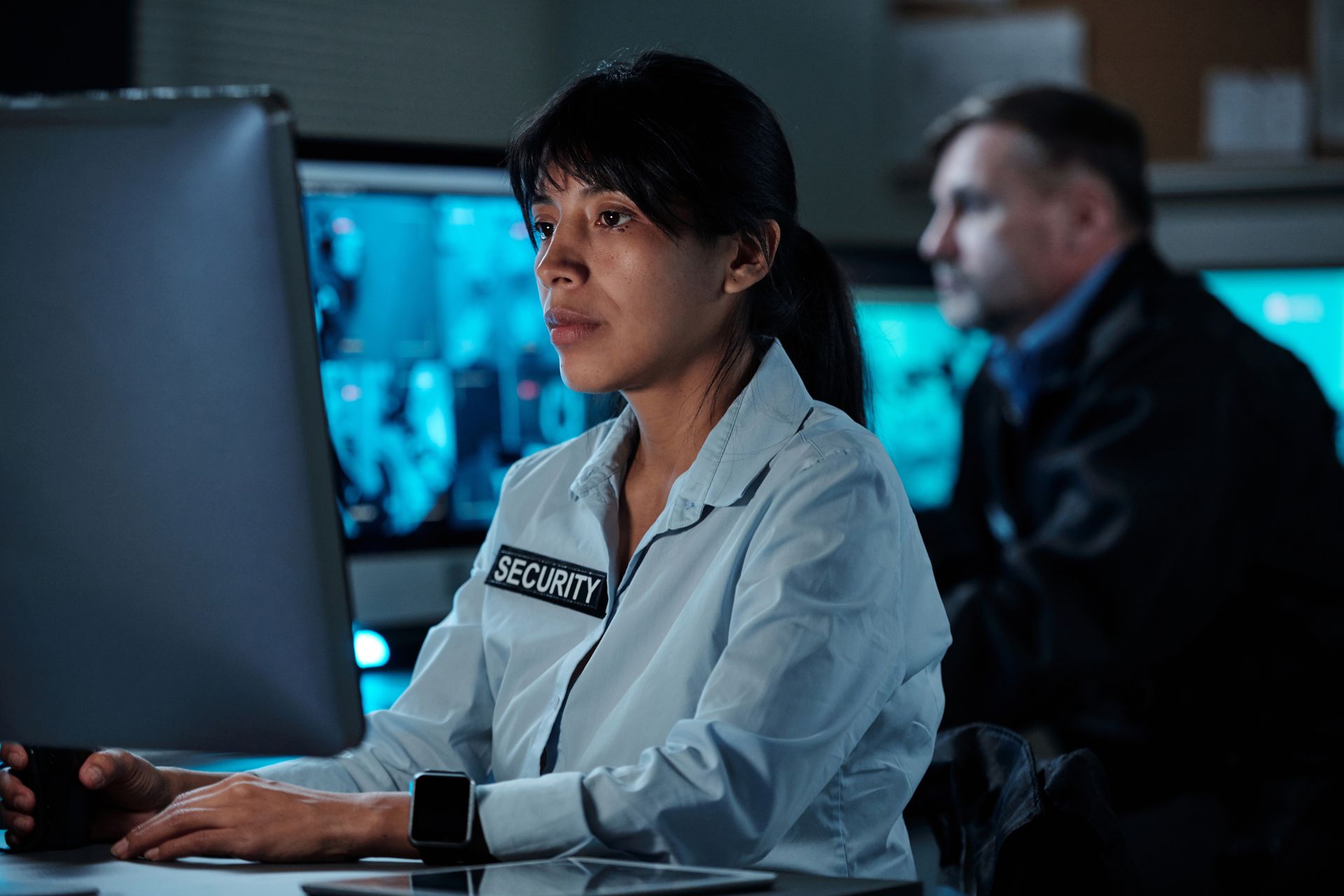 Woman in security uniform monitors screens with a man in a dark room.