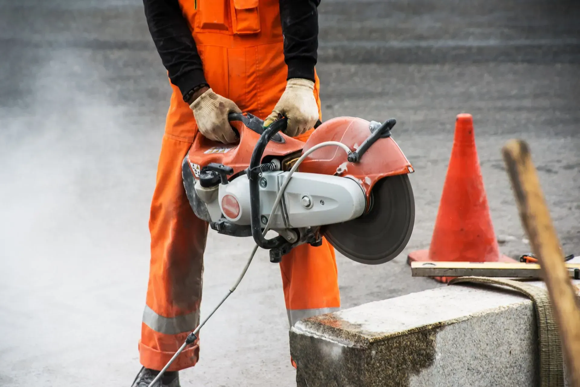 A construction worker in orange overalls uses a circular concrete saw, creating dust near an orange traffic cone.