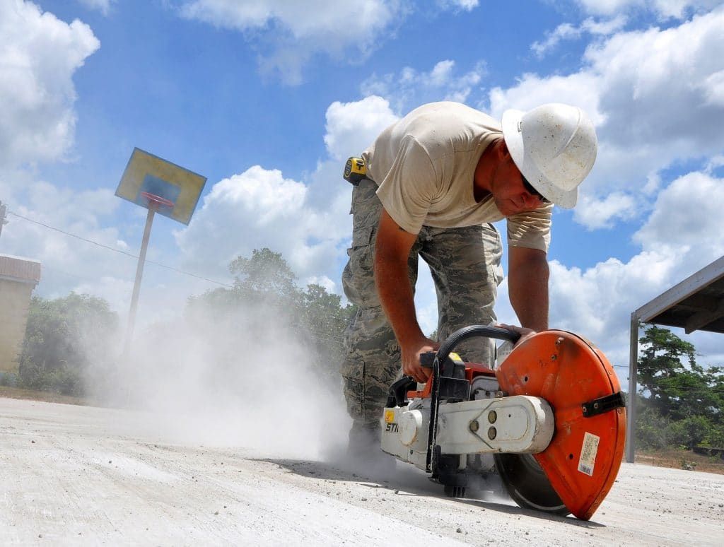 Construction worker using a concrete saw, creating dust. Outdoors, under a blue sky, near a basketball hoop.