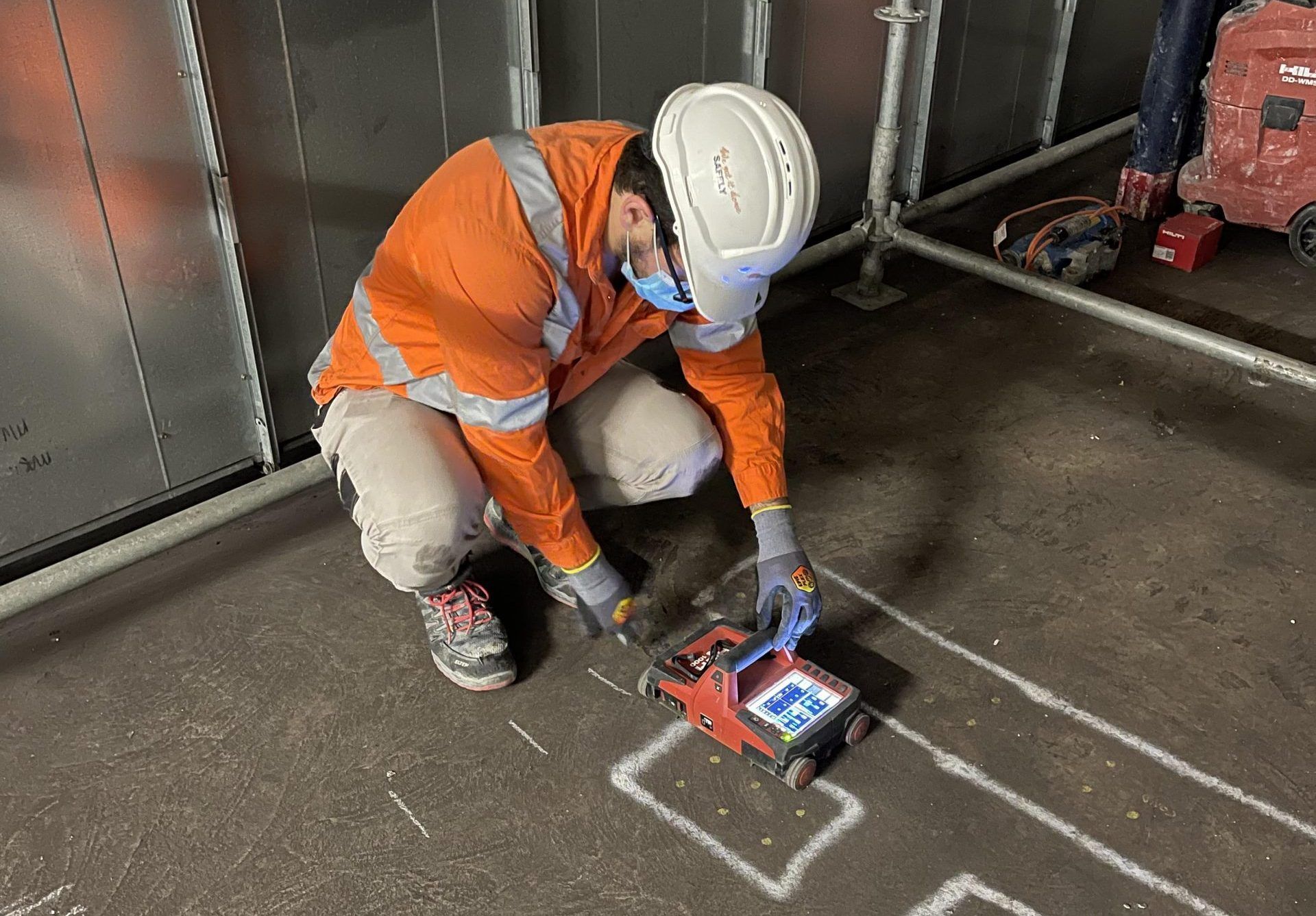 Construction worker in orange vest and hard hat using a red device on a floor.