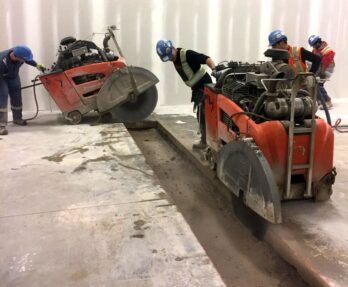 Construction workers in hard hats use walk-behind concrete saws to cut a trench in an indoor concrete floor.