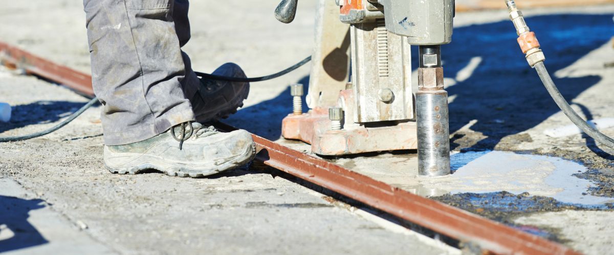 A worker drills a hole in concrete. The worker wears work boots and pants. A metal bar is on the concrete.