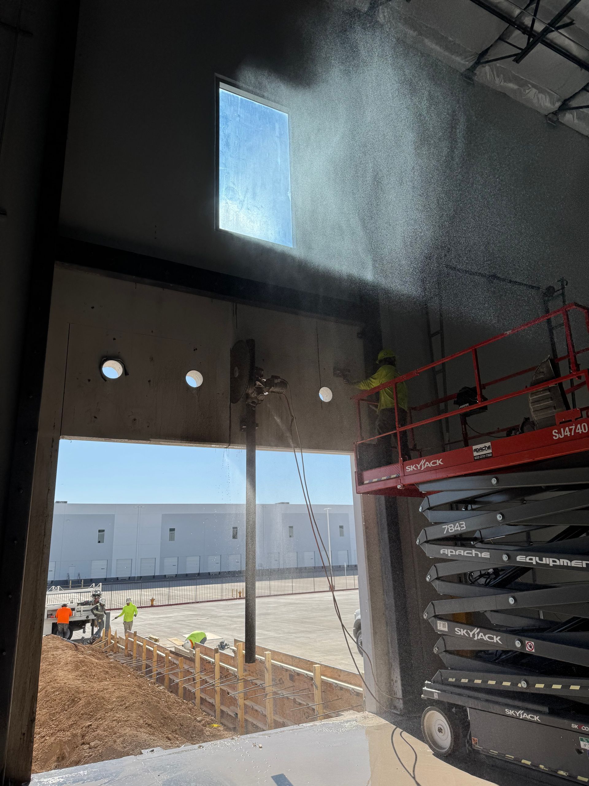Construction worker cutting concrete with a saw, spraying water. A scissor lift and a warehouse are visible.
