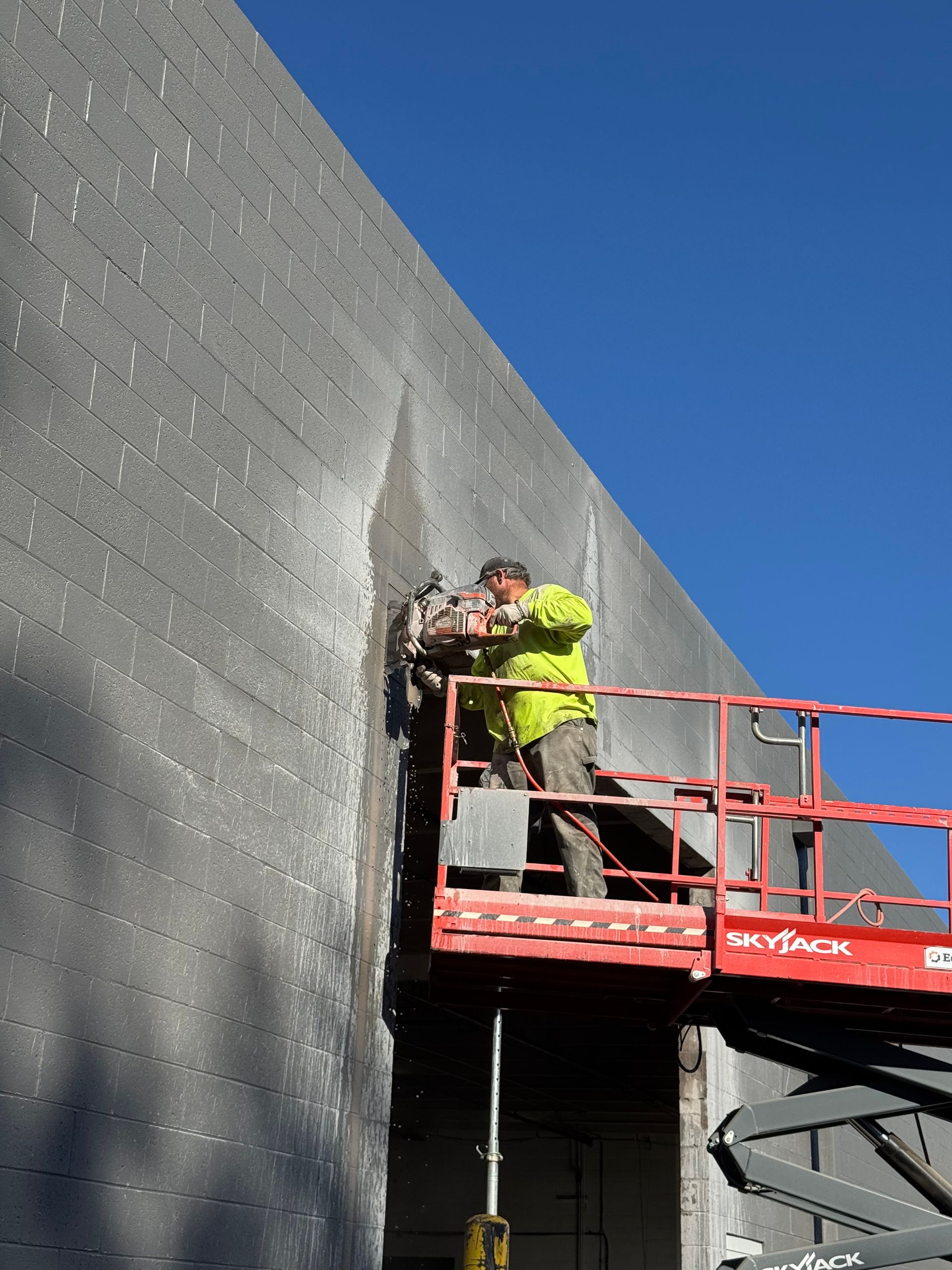 Construction worker using a saw to cut concrete on a building exterior, wearing safety gear, outdoors.