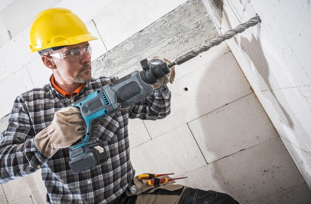 Construction worker drilling into a concrete wall with a power drill; wearing a yellow hard hat and safety glasses.
