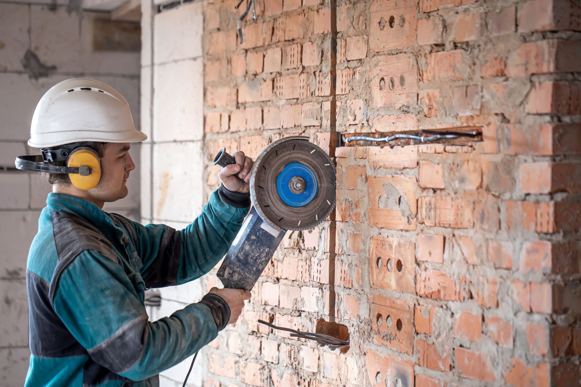 Construction worker cutting brick wall with power tool, wearing safety gear.