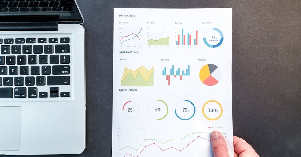 A person is holding a piece of paper with graphs on it in front of a laptop.