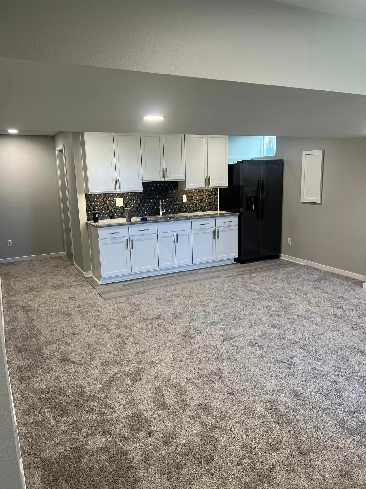 A kitchen in a basement with white cabinets and a black refrigerator.