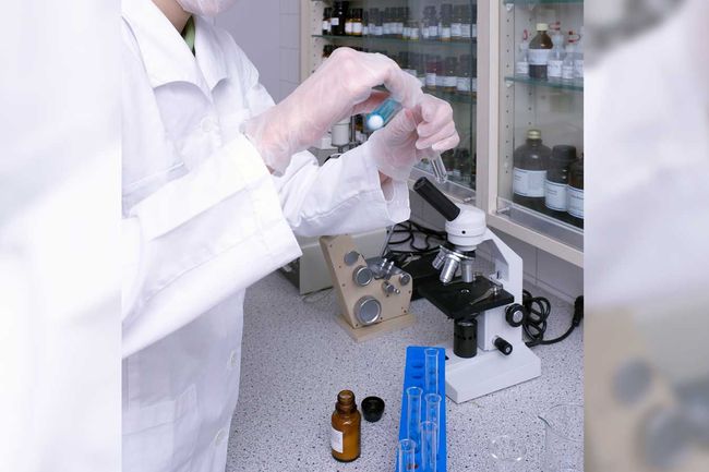Persona con bata y guantes de laboratorio manipulando un tubo cerca de un microscopio en un entorno de laboratorio.
