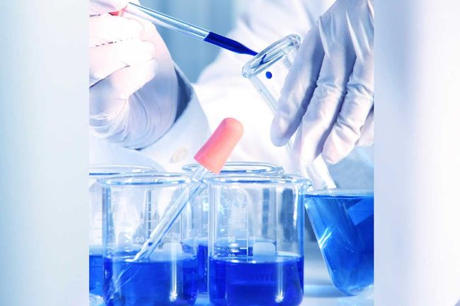 Científico con bata de laboratorio y guantes usando una pipeta para añadir líquido azul a un vaso de precipitados.