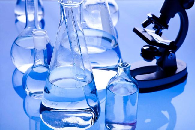 Material de vidrio de laboratorio con líquido en su interior y un microscopio sobre una superficie azul.