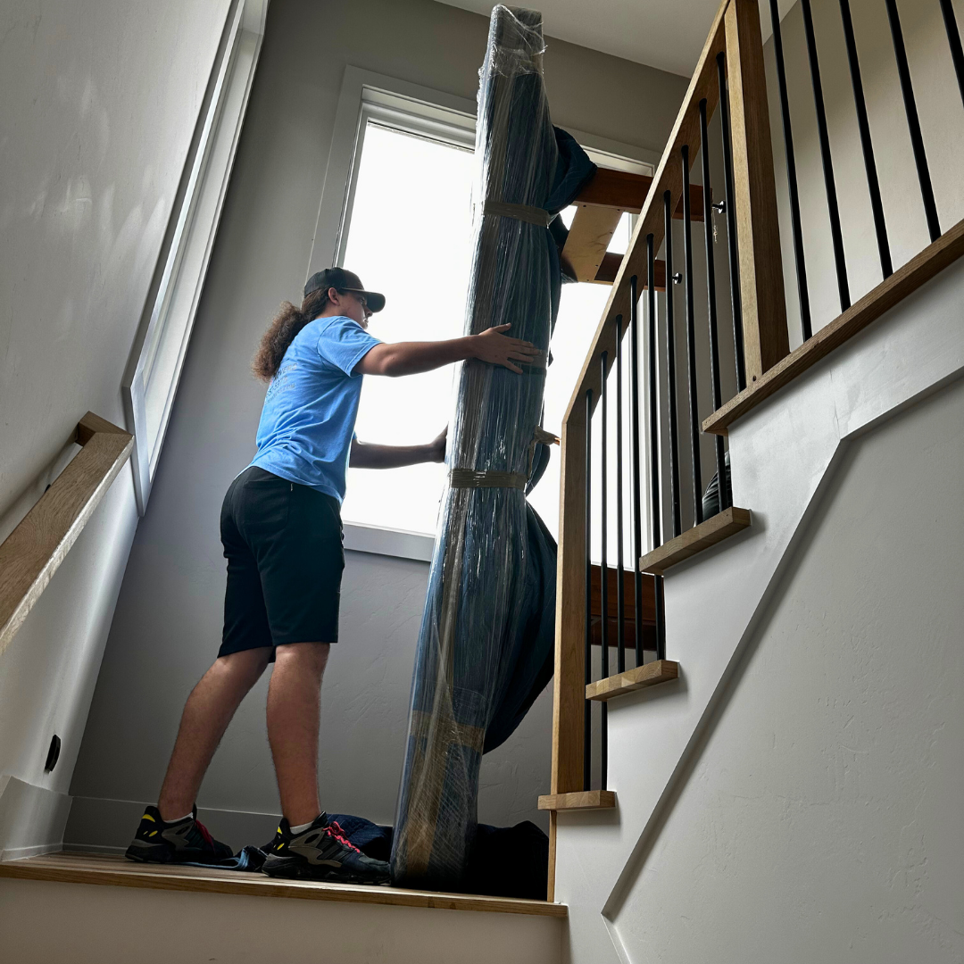 Person carrying a large, rolled-up rug up a staircase.