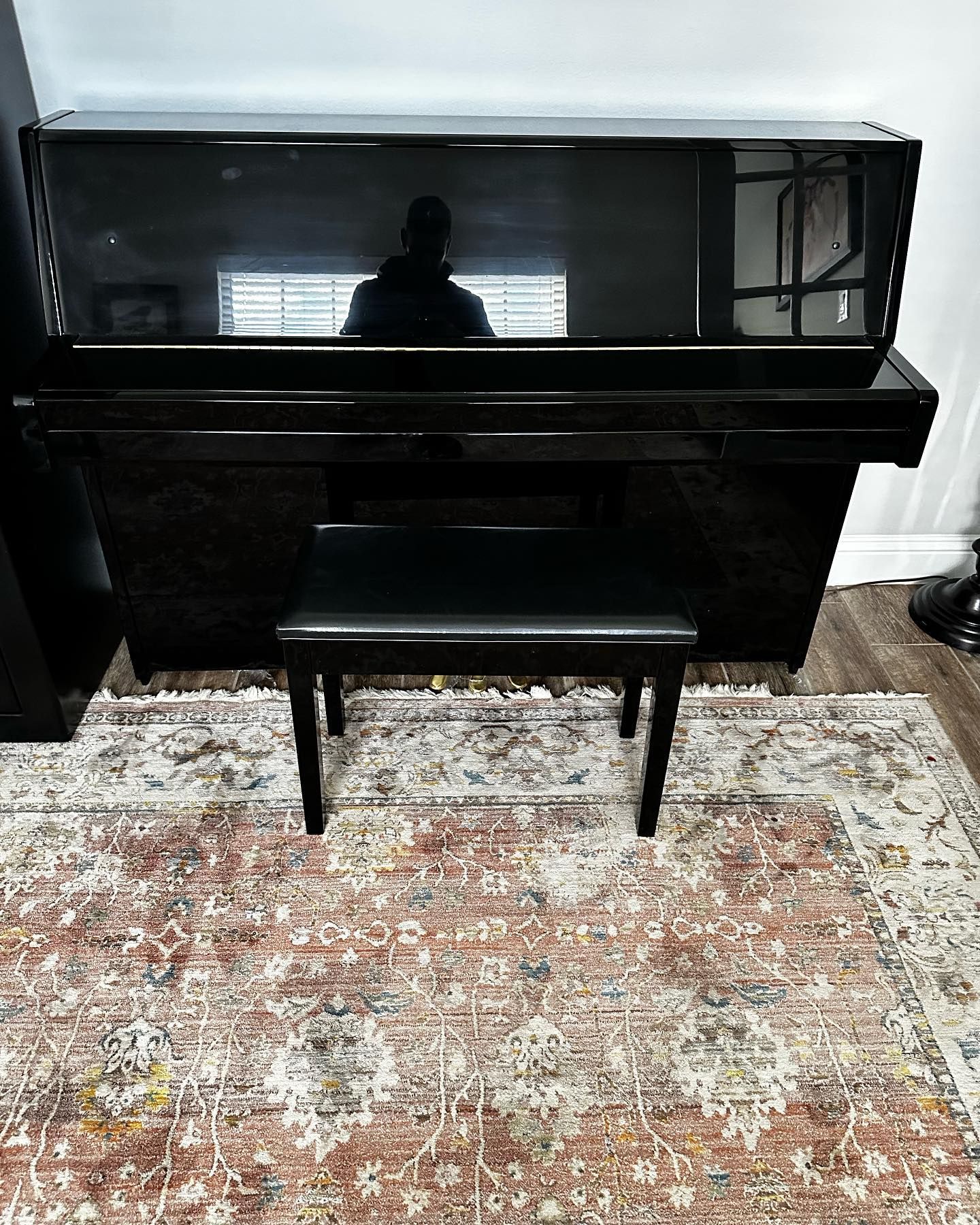 Black piano with a bench on a patterned rug; person reflected in the piano's surface.