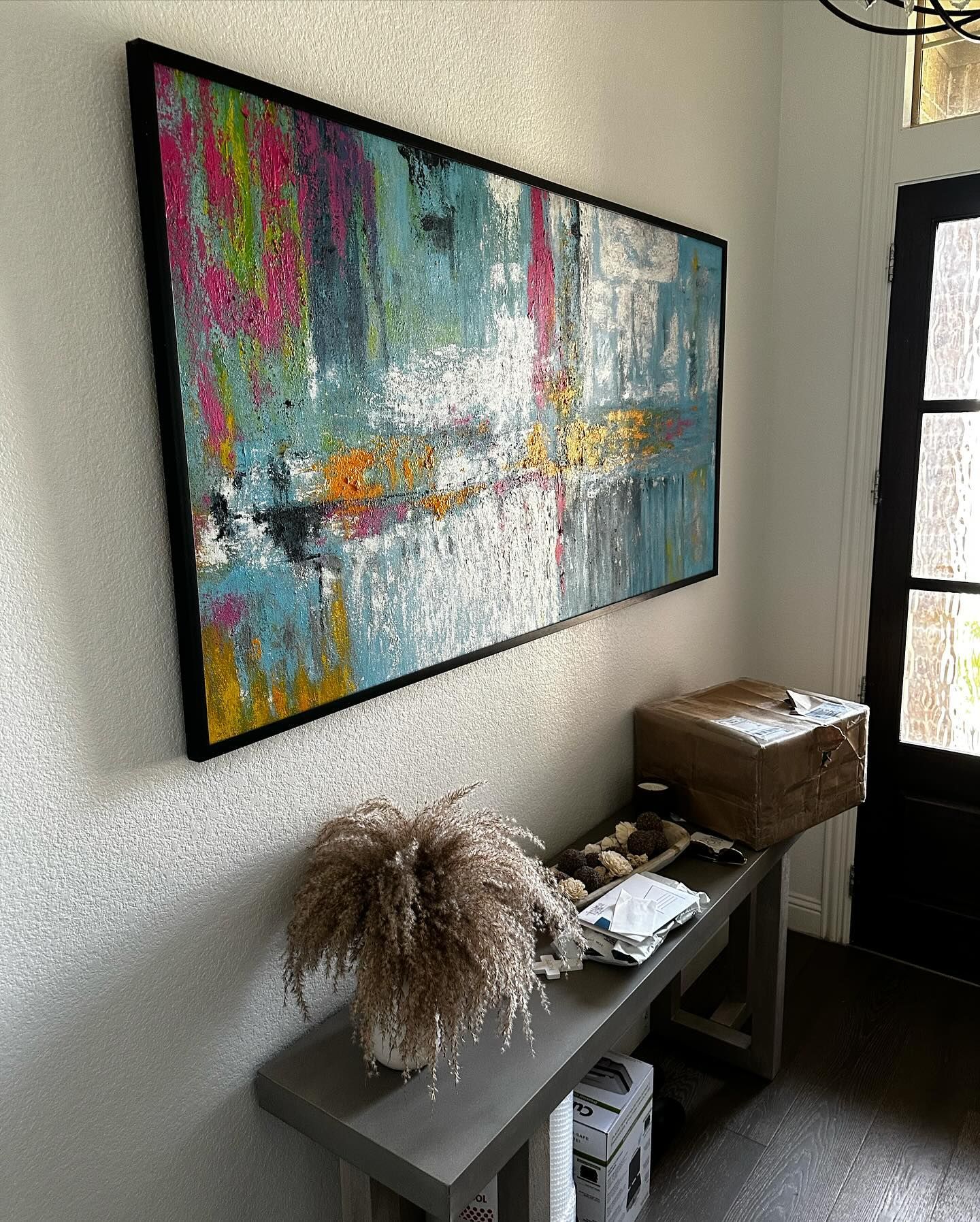 Abstract art with pink, blue, and yellow colors hangs above a gray bench with decor and a doorway.