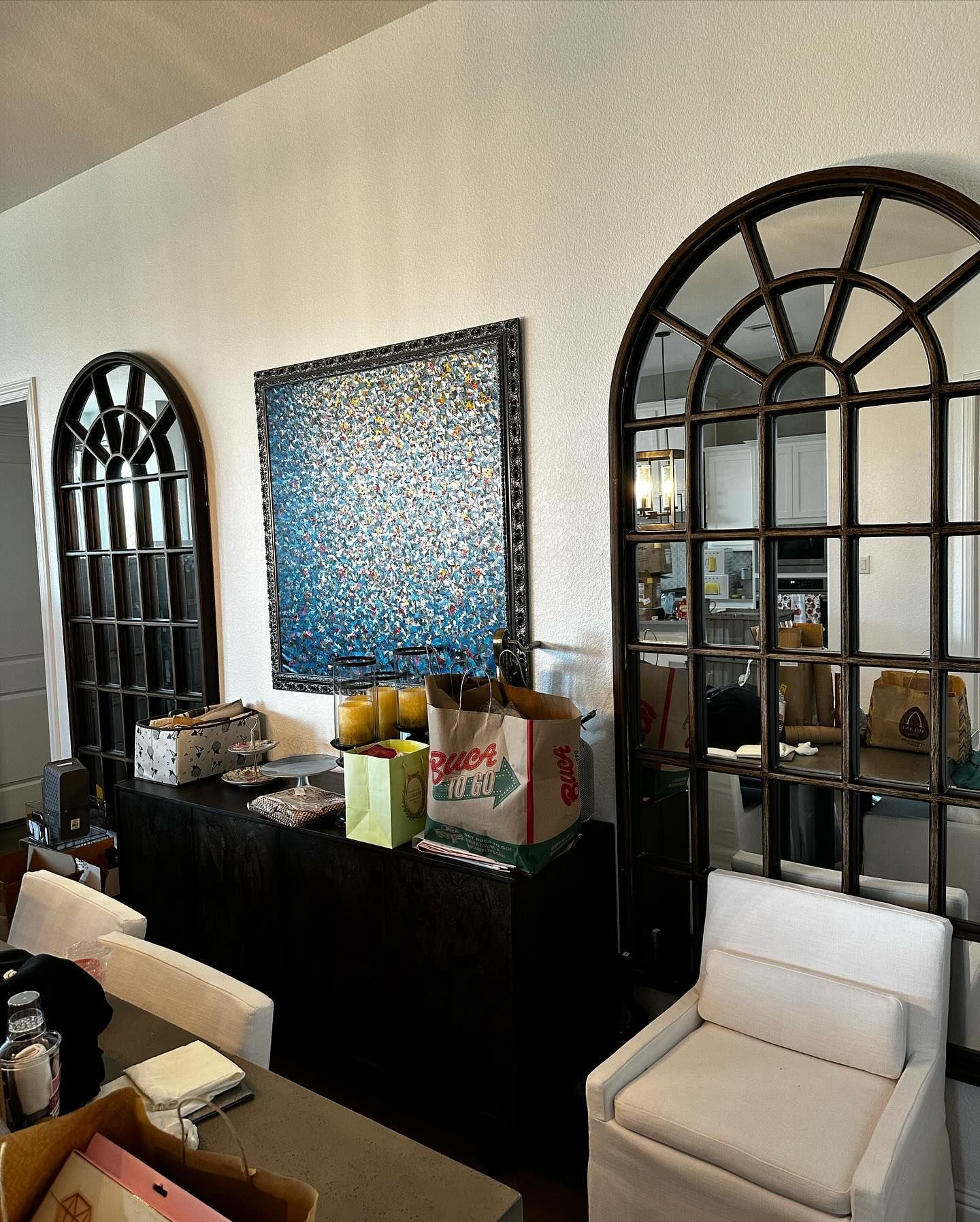Two arched mirrors flank a mosaic art piece above a dark cabinet. A white chair and dining table are in the foreground.
