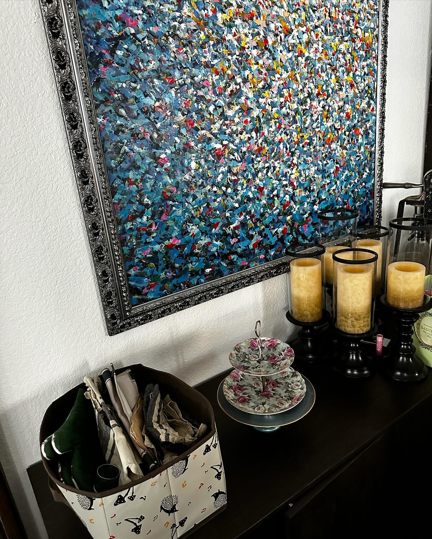 Abstract blue painting above a dark cabinet with candles, tiered plates, and a decorative box.