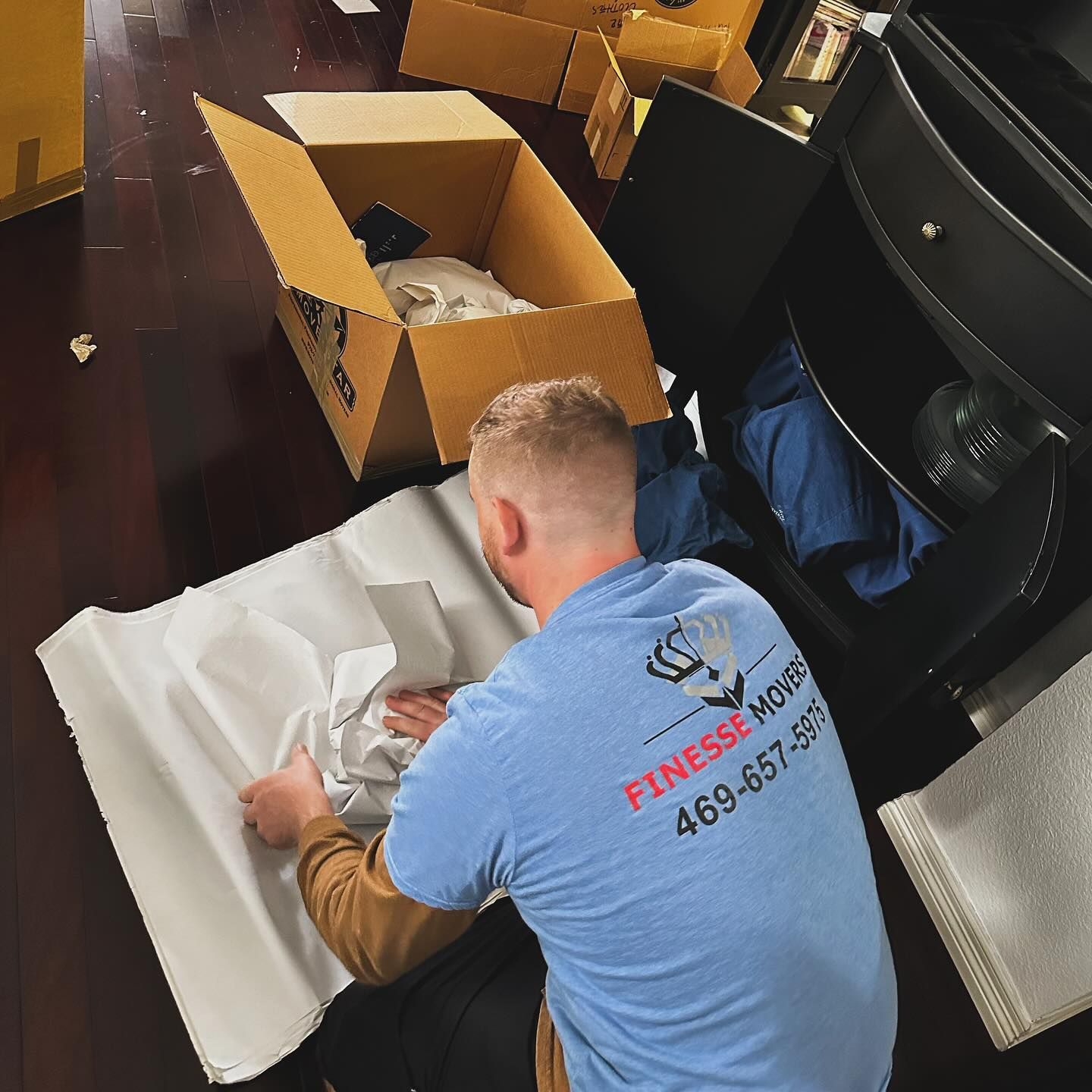 Man in blue shirt wrapping items for moving, near boxes and furniture.