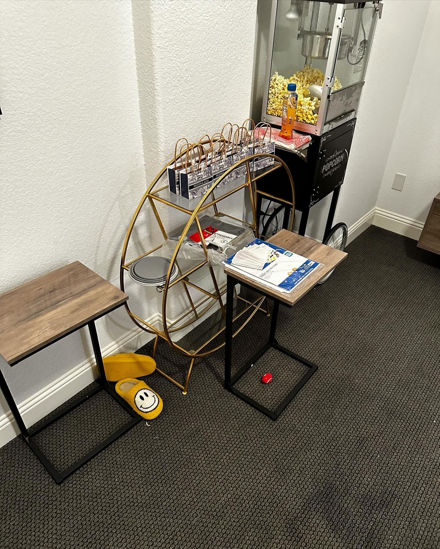 A gold-colored Ferris wheel display holding various snacks, next to a popcorn machine and two small tables.