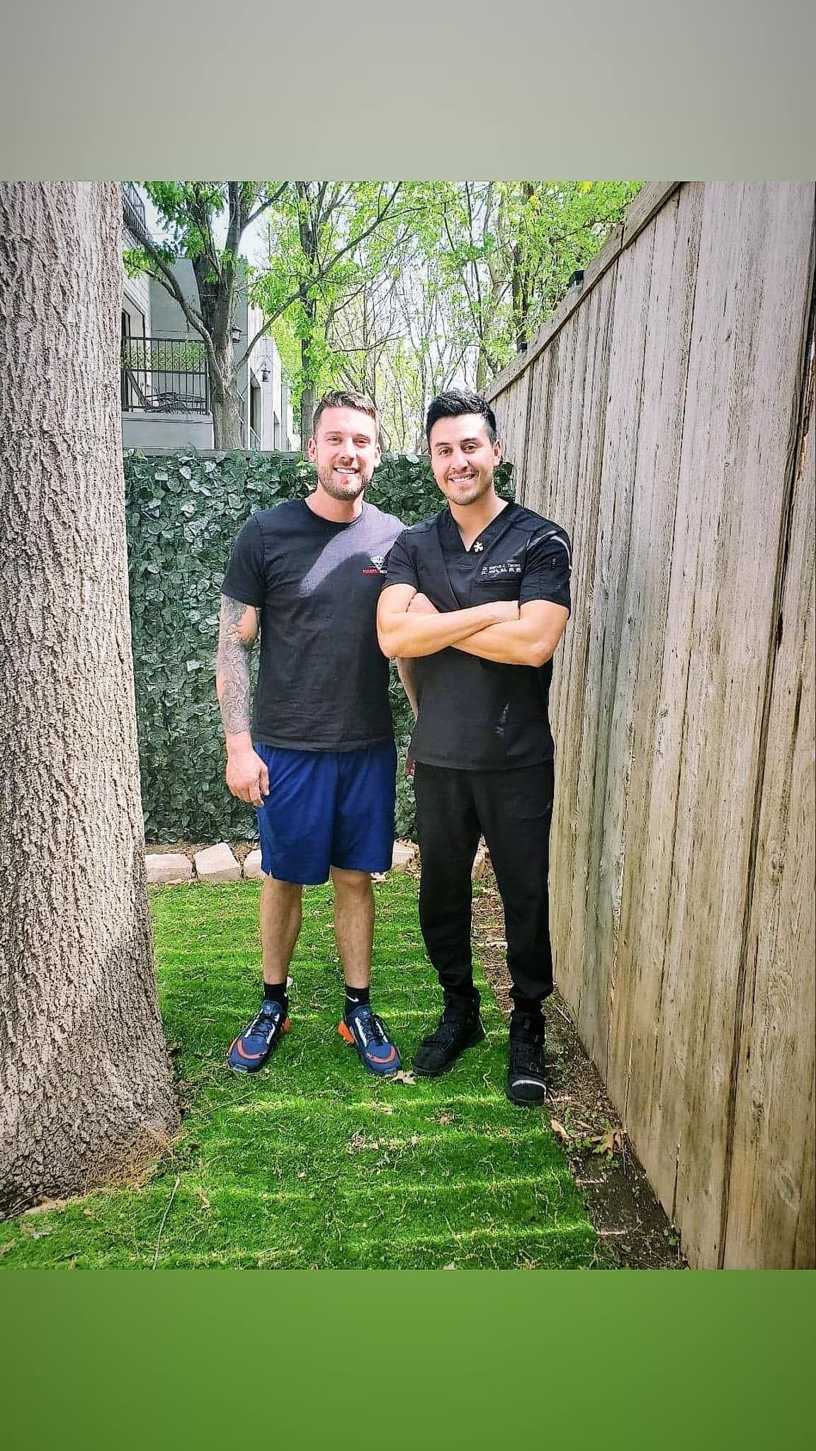 Two men standing outside, one with tattoos wearing shorts and a t-shirt, the other in black scrubs.