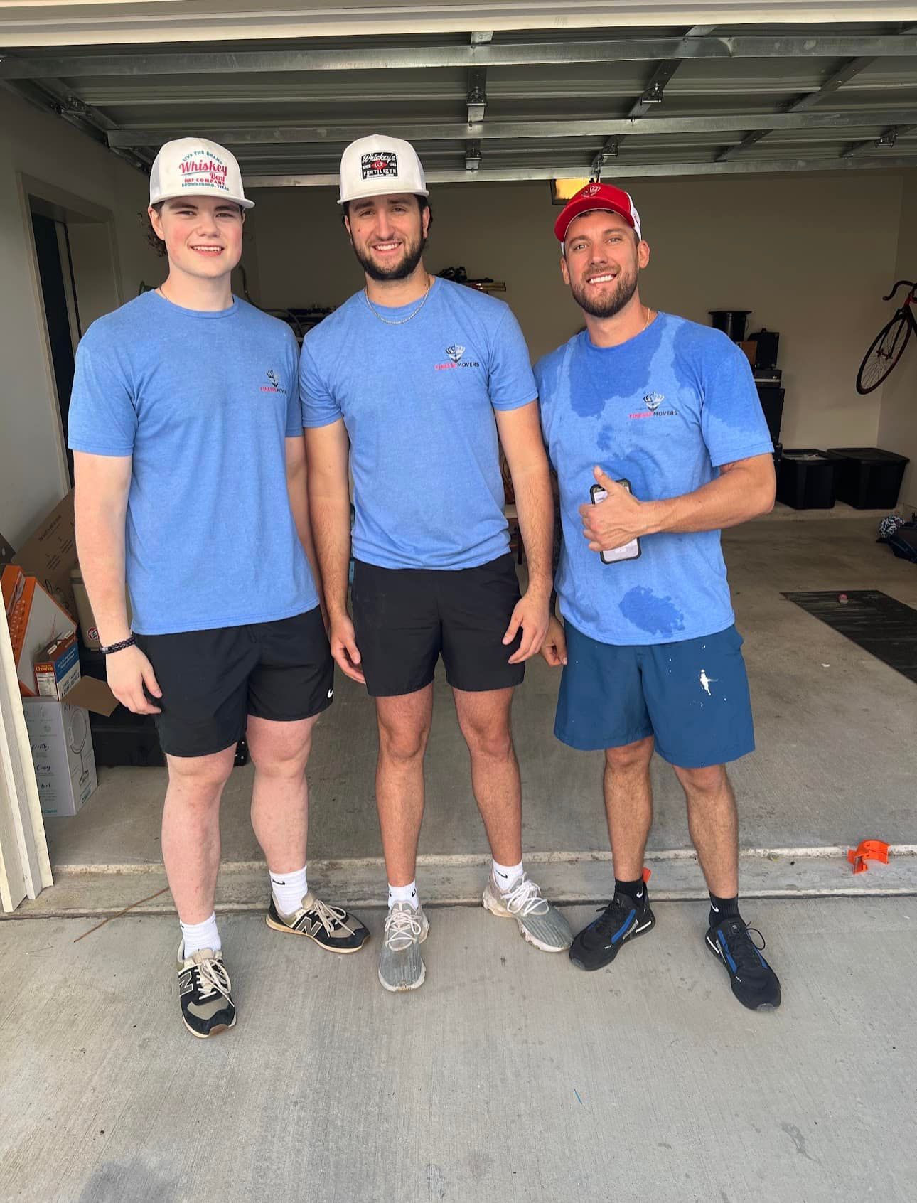 Three men in blue shirts and shorts stand in a garage. Two wear hats. One holds a water bottle.