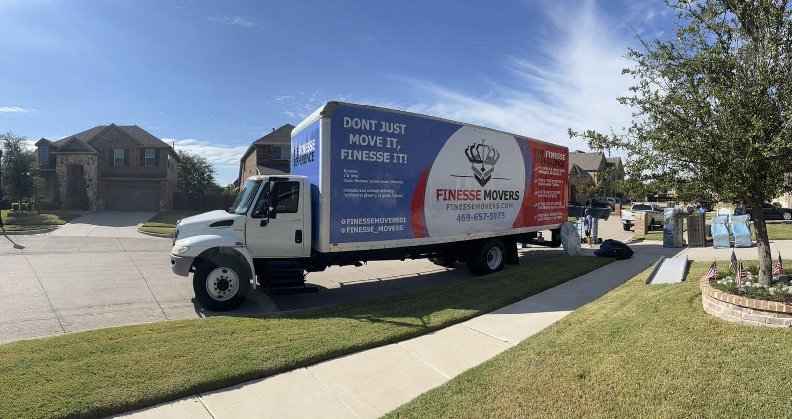 Moving truck parked on a residential street. Boxes and house in the background. Blue sky.