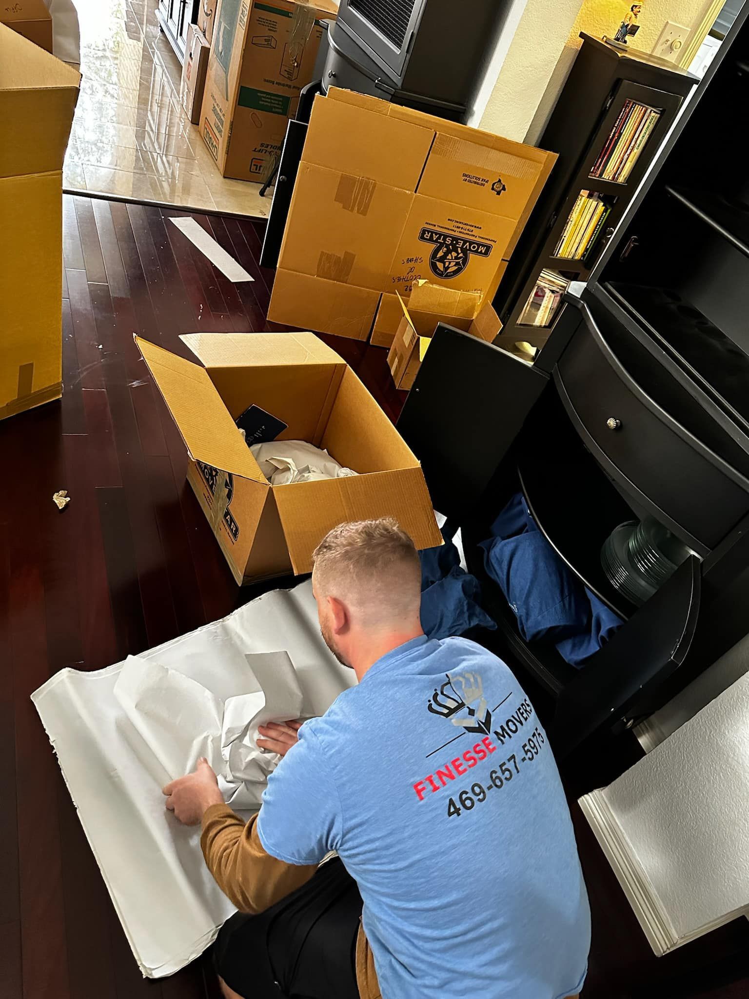 Man in blue shirt packing items, surrounded by boxes and furniture.