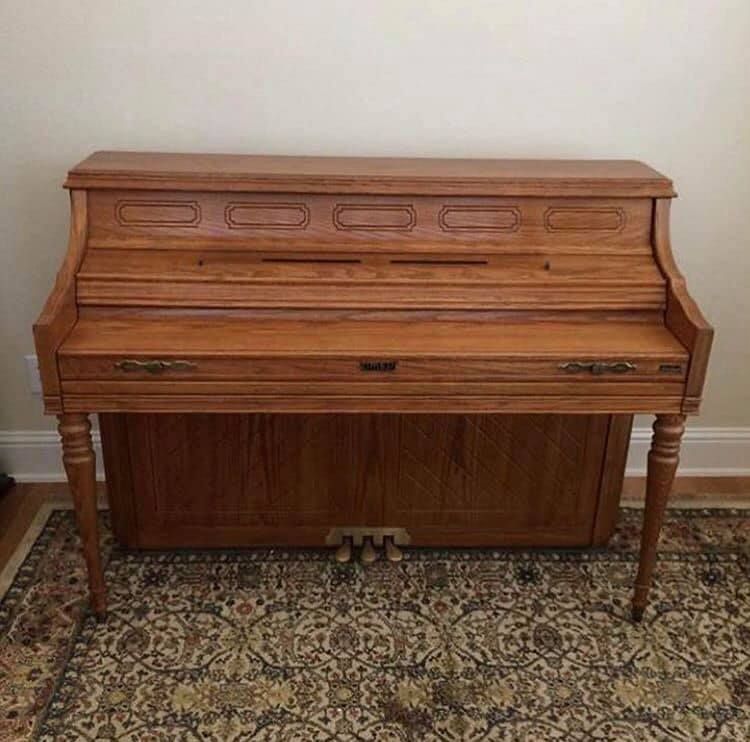 Wooden upright piano on a patterned rug; light-colored walls in the background.