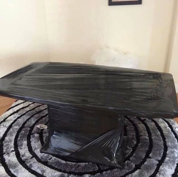Black table wrapped in plastic on a circular gray and white rug, in a room with light walls and wood floors.