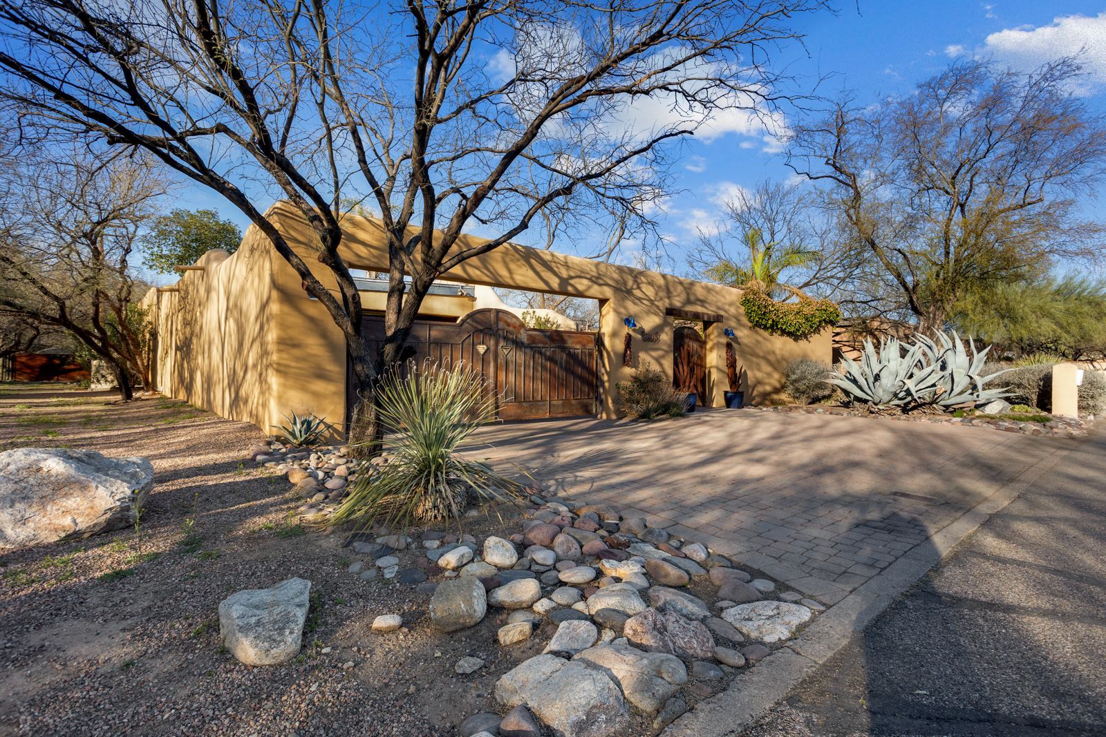 Desert home with stucco walls, large wooden gate, and gravel driveway under a blue sky.