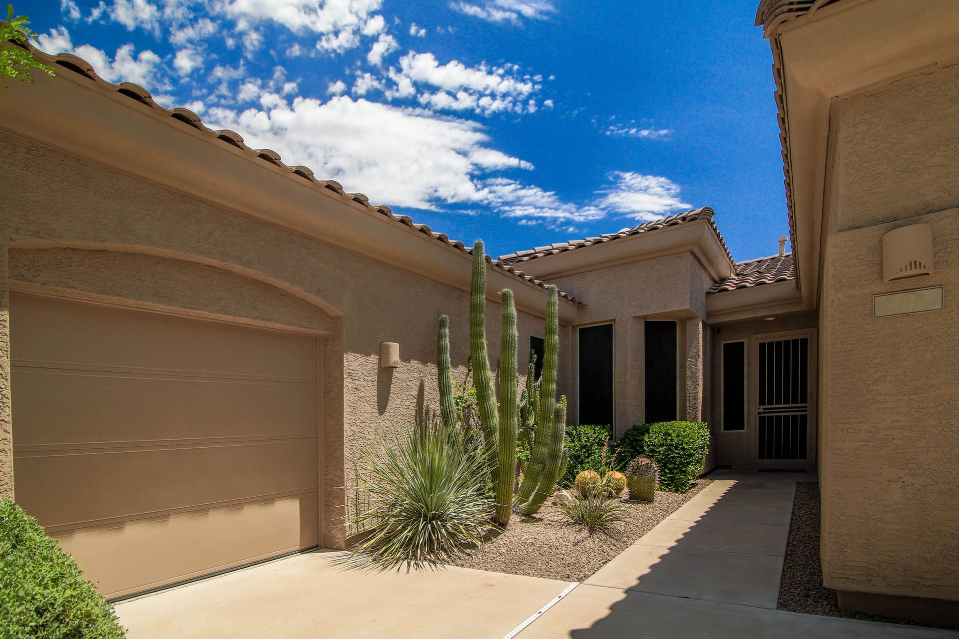 Tan stucco house exterior with a garage, front door, and desert plants under a bright blue sky.