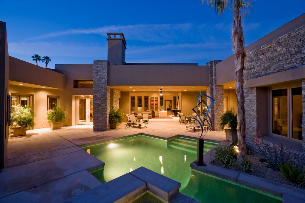 A modern home's courtyard with a pool glowing at night; tan stucco walls, palm tree, patio furniture.